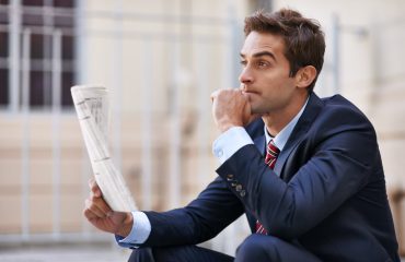 A well-dressed person holding a newspaper looks into the distance with a contemplative expression.